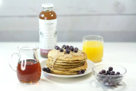 vegan pancakes next to a bottle of maple syrup, orange juice, blueberries and thrive remedies