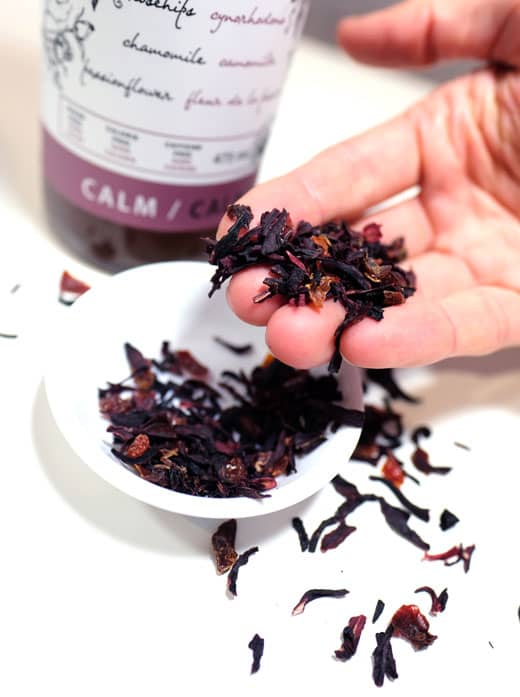 woman's hand holding hibiscus tea above a white dish with a bottle of thrive remedies calm tea in the background